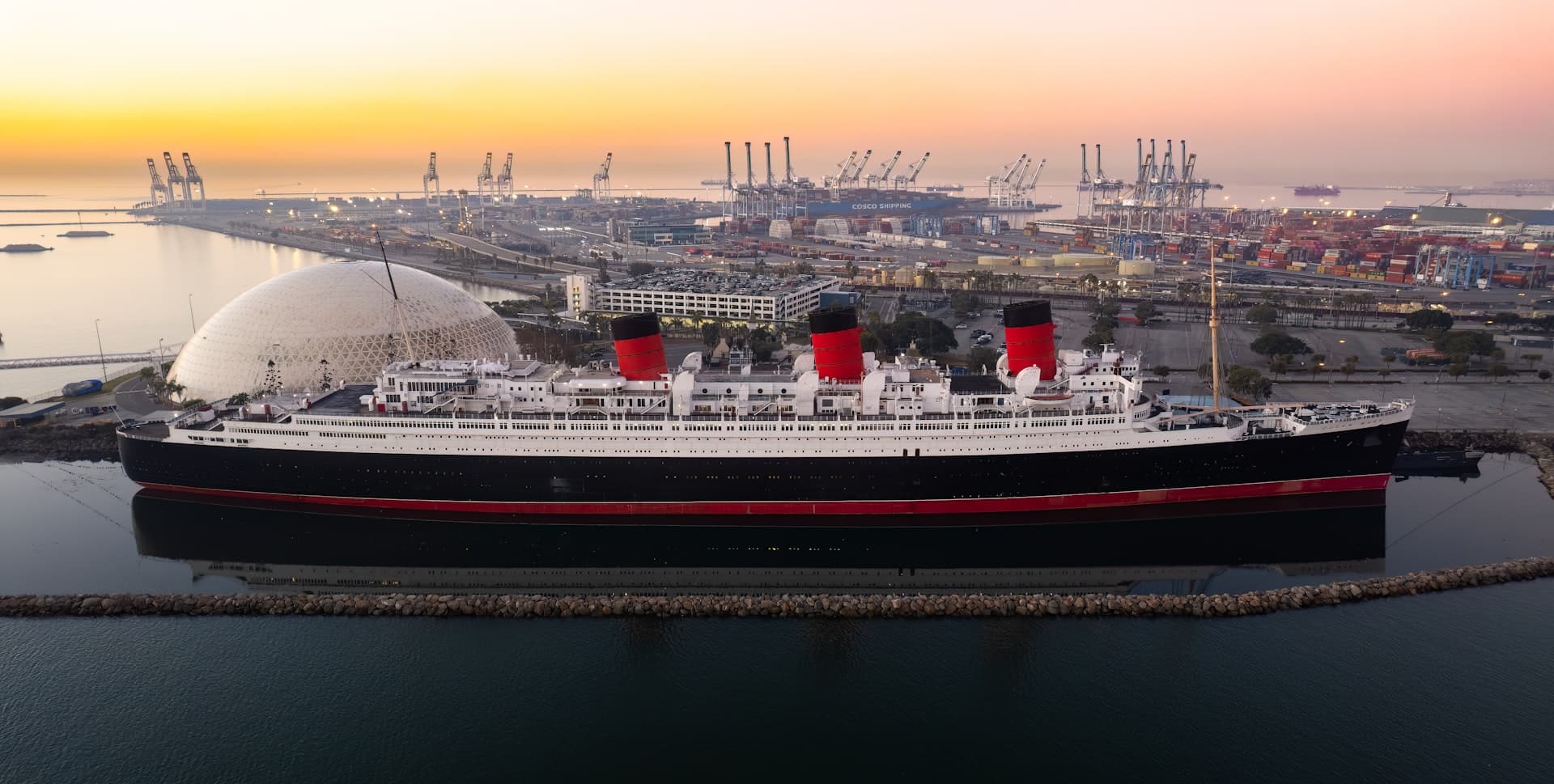 Queen Mary ship docked in Long Beach harbor at sunrise with port in background